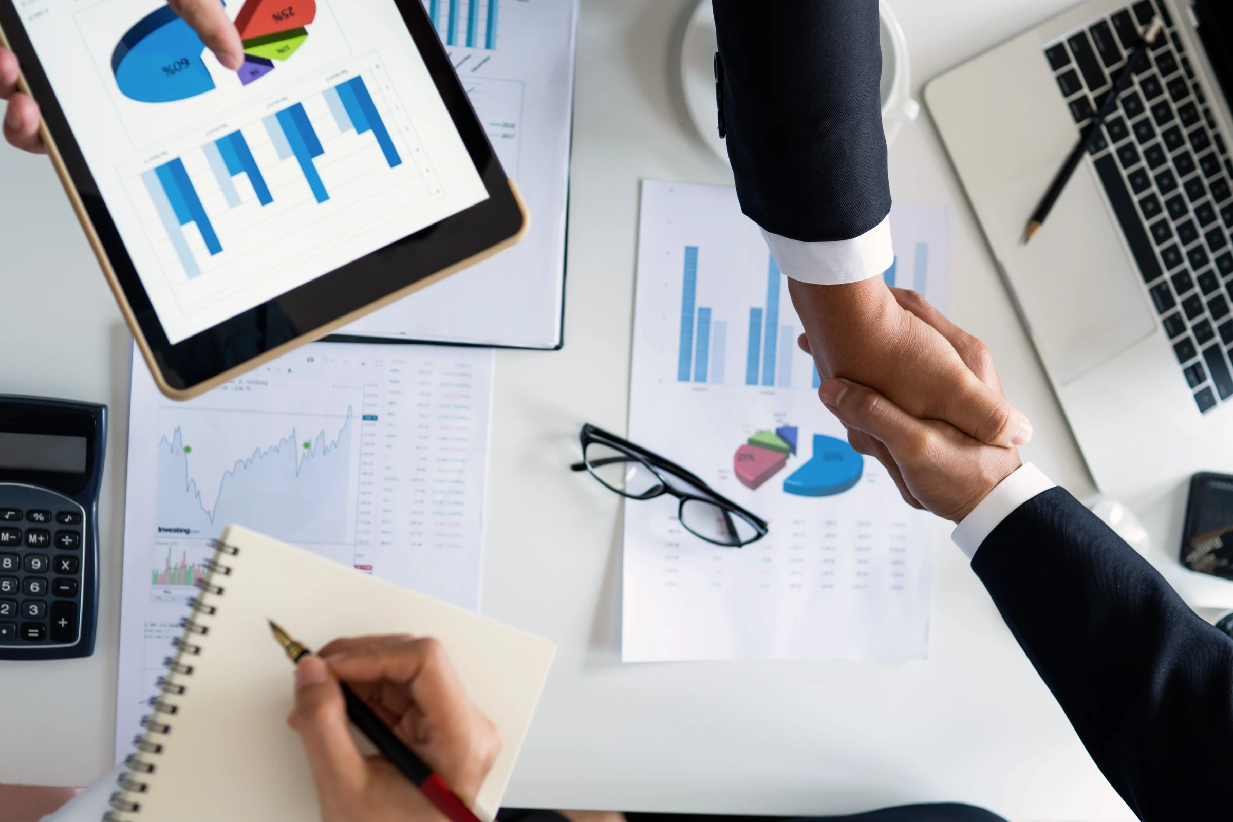 Business partners shaking hands over charts, documents, a laptop and a tablet.